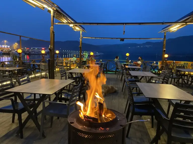 Mavi saatte deniz kıyısındaki ahşap teras, ateş çukuru ve ışıklar